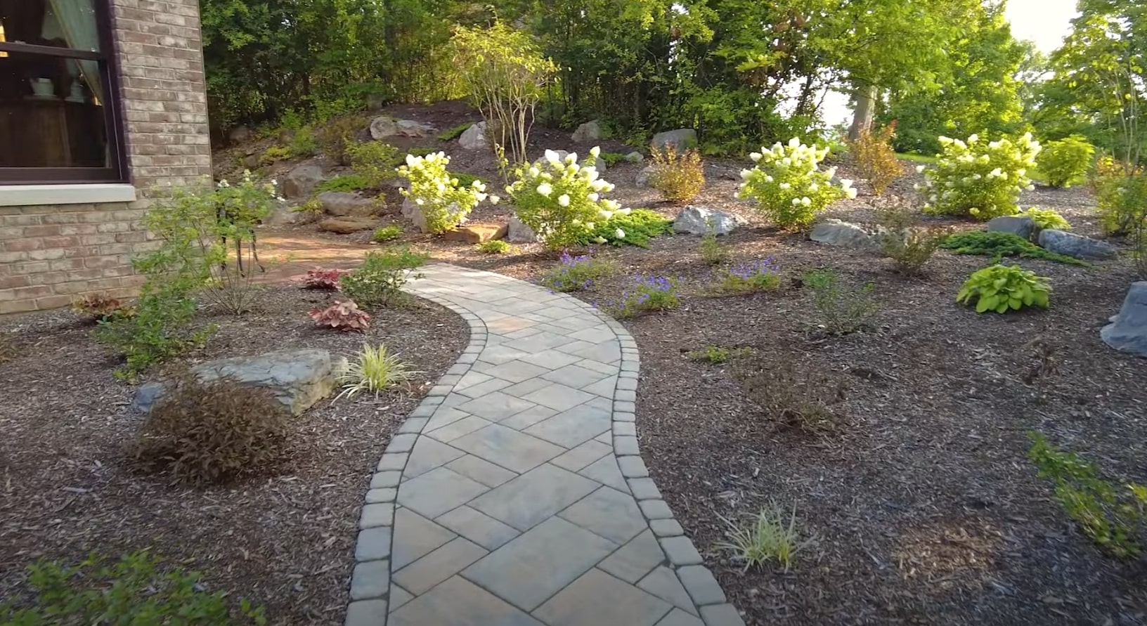 Hardscape Walkway Installation with Professional Landscape Design Curved stone paver walkway through professionally landscaped garden beds with flowering shrubs, decorative rocks, and mature plantings